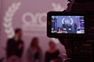 Ein Kamerabildschirm, auf dem man drei Personen und das Logo des Arc-Filmfestivals sieht. Im Hintergrund sieht man das gleiche Bild in groß und verschwommen. 