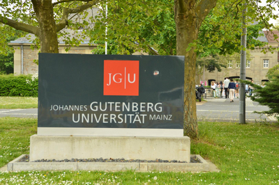 Ein Schild mit dem JGU-Logo und der Aufschrift "Johannes Gutenberg-Universität Mainz". Darunter und dahinter Wiese und Bäume. Im Hintergrund der Eingang zur Universität Mainz. 