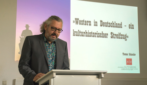 Thomas Schneider. Dahinter eine Powerpoint Folie: "Western in Deutschland - ein kulturhistorischer Streifzug" ©Filmz