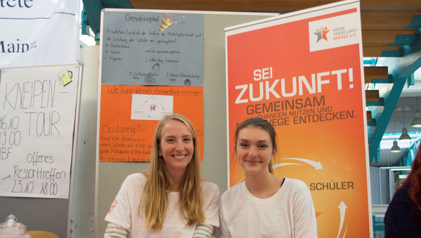 Zwei Studierende sitzen vor dem Banner der Hochschulgruppe "Rock Your Life! Mainz".