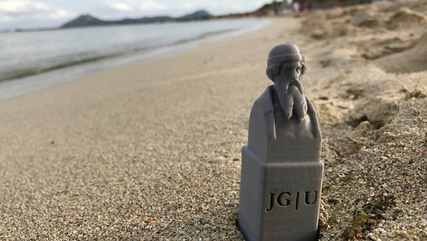 Eine kleiner silberfarbene Gutenberg-Statue mit der Aufschrift JG|U am Strand.