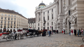 Pferdekutschen und Tourist:innen sind vor der Wiener Hofburg zu sehen.