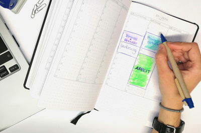 Ein offener Kalender wird im linken Bildrand von einem Laptop gesäumt. Im Kalender sind Zeiten für "Arbeit", "Seminar", "Vorlesung" und "Hausarbeit" farblich ausgeblockt. Darüber liegt eine Hand, die einen Kugelschreiber hält.