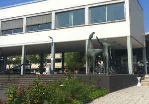 Das Philosophicum der Uni Mainz: Eine reduzierte Statue von einem Mann, der neben einem Pferd steht. Seid Blick geht gen blauer Himmel, danach streckt er auch die Hand aus. Es ist, als reckt er sich nach der feinen Wissenschaft. Davor befindet sich ein grünes, saftiges Blumenbeet. Es ist Sommer.
