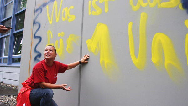 Eine Frau im roten T-Shirt hockt vor einer Gebäudewand, die ein Graffiti aufweist.