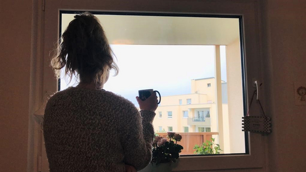 Eine Studentin steht mit dem Rücken zur Kamera am Fenster und blickt nach draußen auf ein Hochhaus vor bedecktem Himmel. In der Hand hält sie eine Tasse.