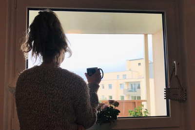Eine Studentin steht mit dem Rücken zur Kamera am Fenster und blickt nach draußen auf ein Hochhaus vor bedecktem Himmel. In der Hand hält sie eine Tasse.