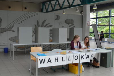 Zwei Frauen sitzen hinter einem Tisch. An dem Tisch steht "Wahllokal". Im Hintergrund sind Wahllokale zu sehen.