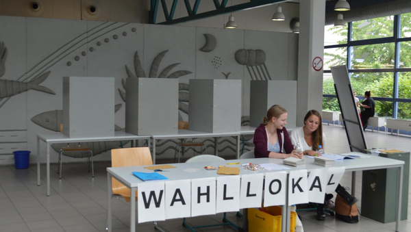 Zwei Frauen sitzen hinter einem Tisch. An dem Tisch steht "Wahllokal". Im Hintergrund sind Wahllokale zu sehen.