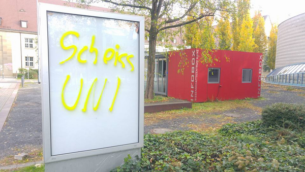 Eine leere Schautafel, auf die in gelber Schrift "Scheiss Uni" gesprüht wurde. Im Hintergrund Wiese und Gebäude.