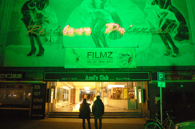 Ein Eingang zu einem Kino. Die Wand ist grün angestrahlt, auf der Wand ist eine rosafarbene Leuchtschrift sowie ein FILMZ-Plakat. Darunter ist ein Durchgang. Zwei Menschen gehen auf den Eingang zu. 