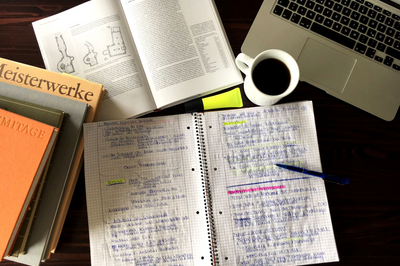Ein beschriebener Block, ein Stift, ein offenes Buch und ein Bücherstapel, eine Tasse Kaffee und ein Laptop auf einem Tisch.