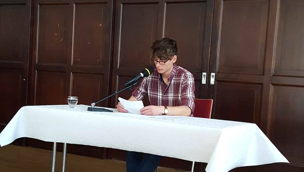 Ein junger Mann mit Brille sitzt an einem Tisch und liest von einem Blatt ab.