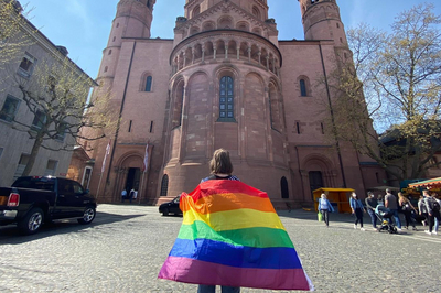 Vor dem Mainzer Dom ist eine Studentin von hinten zu sehen. Auf dem Rücken trägt sie eine Regenbogenflagge. Um sie herum laufen Passant:innen über den Domplatz.