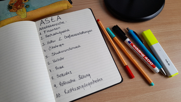 In einem aufgeschlagenen Notizbuch sind die Arbeitsbereiche des AStA niedergeschrieben. Ringsum liegen Stifte verteilt.