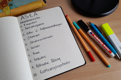 In einem aufgeschlagenen Notizbuch sind die Arbeitsbereiche des AStA niedergeschrieben. Ringsum liegen Stifte verteilt.