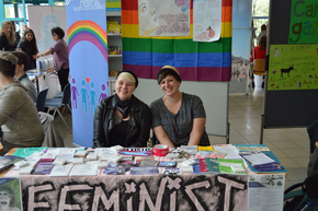 Zwei Frauen sitzen an einem Messestand mit der Beschriftung "Feminist".