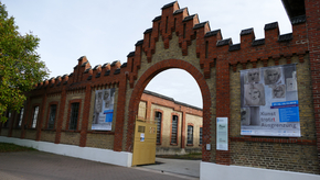 Das Bild zeigt den Haupteingang des damaligen Konzentrationslagers und der heutigen Gedenkstätte in Osthofen: Das hohe Eingangstor überragt einen großen Baum zur linken Seite, es wird zudem auf beiden Seiten gesäumt von zahlreichen Plakaten, die unter anderem auf eine Kunstausstellung auf dem Gelände hinweisen.