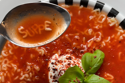 Das Artikelfoto zeigt einen Löffel voll Tomatensuppe über einem Teller. Mit Buchstabennudeln geschrieben steht das Wort Suppe in dem Löffel. Die Suppe ist mit Joghurt, Chiliflocken und Basilikum verziert.