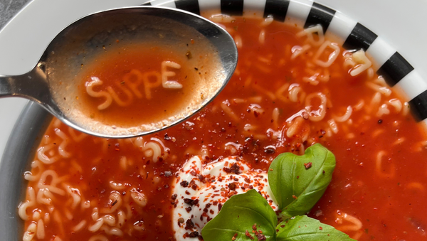 Das Artikelfoto zeigt einen Löffel voll Tomatensuppe über einem Teller. Mit Buchstabennudeln geschrieben steht das Wort Suppe in dem Löffel. Die Suppe ist mit Joghurt, Chiliflocken und Basilikum verziert.