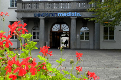 Im Vordergrund Blumen, im Hintergrund ist der Haupteingang der Universitätsmedizin zu sehen.