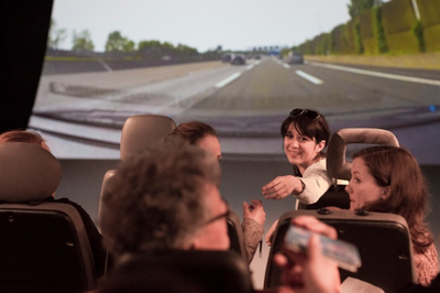 Junge Menschen auf Autositzen. Ganz vorne sitzt eine Frau und dreht sich um. Die Teilnehmer sitzen einer großen Leinwand gegenüber, die eine Autobahn zeigt. 
