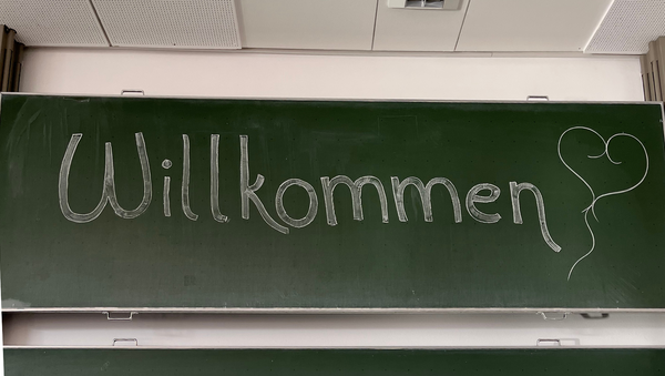 Das Artikelfoto zeigt eine grüne Tafel auf der mit weißer Kreide das Wort "Willkommen" steht. Dahinter wurde ein kleines weißes Herz gezeichnet.