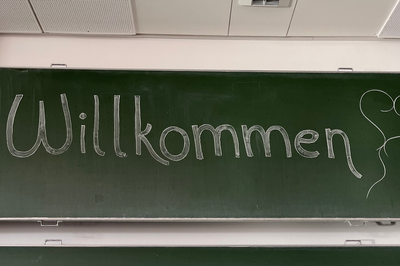 Das Artikelfoto zeigt eine grüne Tafel auf der mit weißer Kreide das Wort "Willkommen" steht. Dahinter wurde ein kleines weißes Herz gezeichnet.