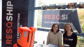 Zwei Studierende sitzen hinter dem Messestand der Hochschulgruppe "RESQSHIP", auf dessen Tisch u.a. eine orange Rettungsweste platziert wurde.