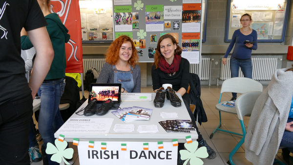 Zwei Vertreter:innen der Irish-Dance-Gruppe sitzen hinter einem Tisch, auf dem Flyer und Schuhe, aber auch ein Laptop liegen, der die Arbeit der Gruppe zeigt. Auf der Stellwand hinter ihnen sind weitere Fotos ihrer Arbeit zu sehen.