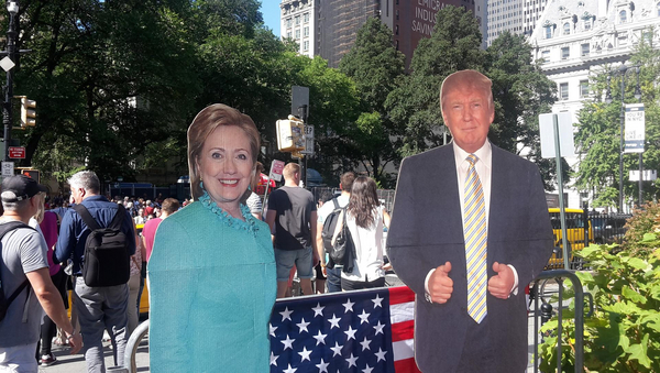 Links eine Pappfigur von Hillary Clinton, rechts Donald Trump. Im Hintergrund Menschen.
