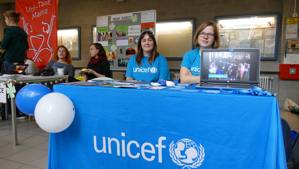 Zwei Mitglieder der Hochschulgruppe von UNICEF sitzen in blauen Shirts hinter dem Messestand, der mit einer blauen Tischdecke versehen ist und auf dem Flyer liegen.