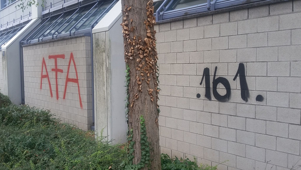 Eine graue Wand, auf die in schwarzer Farbe ".161." und in roter Farbe "AFA" gesprüht wurde. Vor der Wand ein Baum und Wiese.