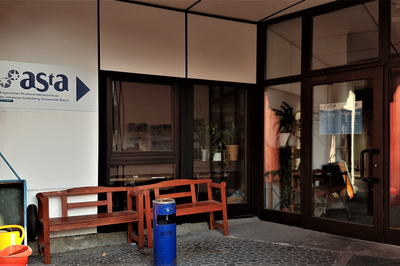 Das Bild zeigt das Büro des AStA im Staudingerweg von außen: Vor der Tür steht eine Bank, darüber und an der Tür selbst ist sein Logo zu sehen.