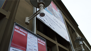 Unter einem großen Plakat an der Alten Mensa, in der die LOB-Tagung beworben wird, steht eine Stellwand mit roten Plakaten und detaillierteren Erläuterungen der Aktion "Wir müssen schließen.".
