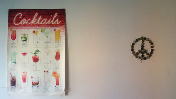 Eine weiße Wand. Links ein Poster mit verschiedenen Cocktail-Sorten. Rechts ein Peacezeichen aus Bierkronen. 