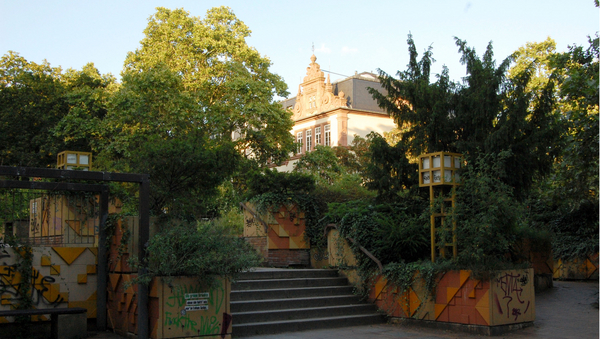 Eine Treppe, umgeben von Mauern, Pflanzen und Bäumen. Im Hintergrund ein Gebäude.