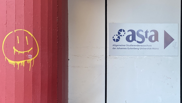Das Bild zeigt den Eingangsbereich des AStA-Sekretariats an der Uni Mainz. Links im Bild ist eine rote Steinsäule zu sehen, auf welcher mit gelber Graffiti-Farbe ein Smiley gesprüht wurde. Rechts an der Wand befindet sich ein weißes Schild mit dem blauen Logo des AStA. 