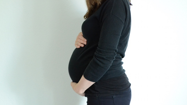 Eine schwangere Frau, dunkel gekleidet, die seitlich vor einer weißen Wand steht und sich ihren Babybauch hält. 