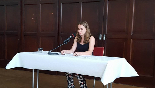 Eine junge Frau sitzt an einem Tisch mit einem Mikrofon und liest von einem Blatt ab.