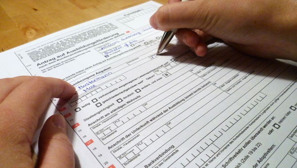Eine Person füllt einen Antrag aus. Man sieht ein Formular auf einem Tisch und zwei Hände. Eine Hand füllt mit einem Kugelschreiber den Antrag aus.