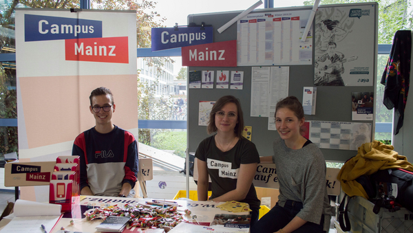 Drei Studierende sitzen hinter einem Tisch mit Flyern. Dahinter befindet sich eine Stellwand mit Campus Mainz-Wandkalendern.