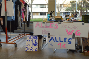 ein sehr vollgestellter Stand neben einer Kleiderstange mit Schild "Alles für Deine WG, alles ab 1 Euro"