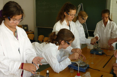 Fünf Mädchen in Kitteln und mit Schutzbrille, die um einen Tisch stehen und chemische Versuche machen.