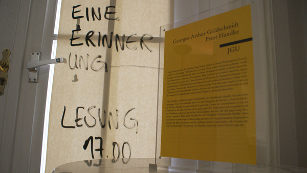 Ein Text auf einem gelben Zettel im Reclam-Stil. Auf eine Glastür geklebt. Daneben steht etwas auf die Glastür geschrieben. 