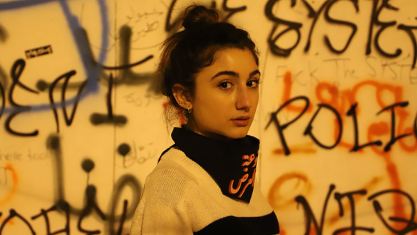 Eine junge Frau steht vor einer Wand voller Graffiti. Ihr Blick sagt vieles, v.a. sieht er in eine ungewisse Zukunft.