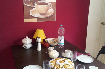 Ein gedeckter Tisch mit Tellern, Gläsern, Kaffeetassen und einem Kuchen in der Mitte. Im Hintergrund eine lila-pinkfarbene Wand mit einem in braun gehaltenem Bild, auf dem zwei Kaffeetassen abgebildet sind und "Cappucino" steht. 