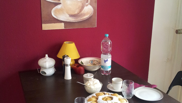 Ein gedeckter Tisch mit Tellern, Gläsern, Kaffeetassen und einem Kuchen in der Mitte. Im Hintergrund eine lila-pinkfarbene Wand mit einem in braun gehaltenem Bild, auf dem zwei Kaffeetassen abgebildet sind und "Cappucino" steht. 