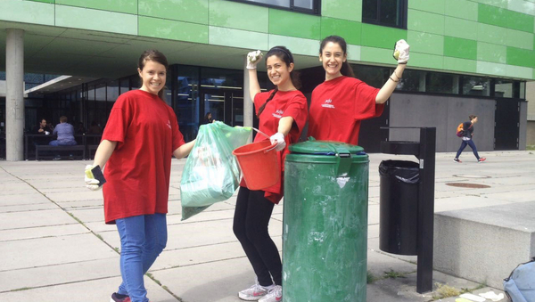 Drei junge Frauen in roten T-Shirts posieren für das Foto. Sie halten einen Mülleimer und eine Mülltüte in der Hand. Im Vordergrund ein grüner Mülleimer, im Hintergrund die grüne Fassade eines Gebäudes.