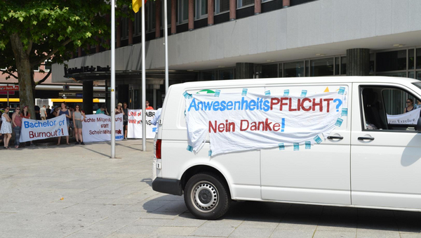 Ein Transporter, an dem ein Laken mit der Aufschrift "AnwesenheitsPFLICHT? Nein Danke!" hängt, steht vor der Rheinland-Pfalz Bank. Im Hintergrund sieht man Demonstrierende ihre Spruchlaken hochhalten.